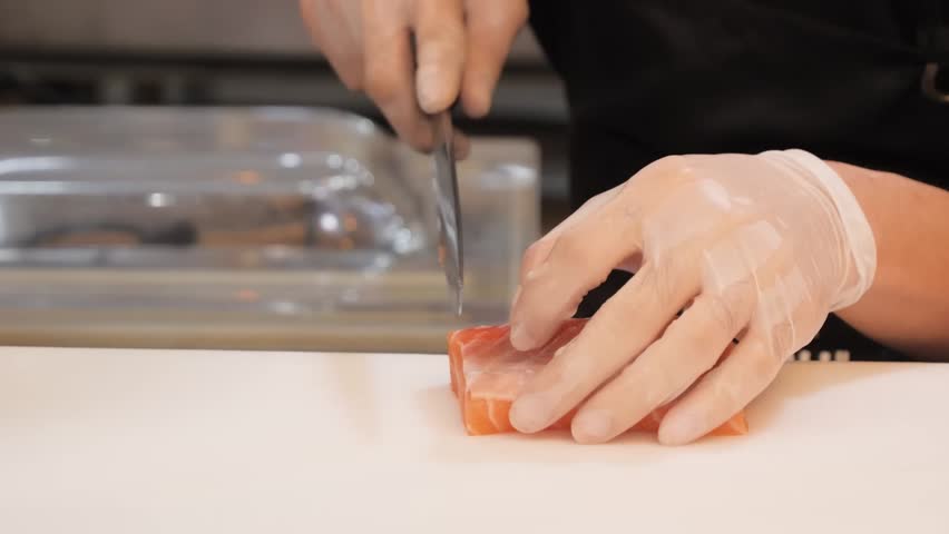 Close-up of a professional sushi chef wearing gloves, skillfully slicing fresh, raw salmon fillet on a white cutting board. The chef uses a sharp traditional Japanese knife to create uniform sashimi p