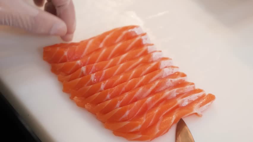 Close-up footage of a professional chef using a sharp traditional knife to precisely slice a fresh, raw Atlantic salmon fillet into delicate sashimi pieces. The vibrant orange meat with white fat marb