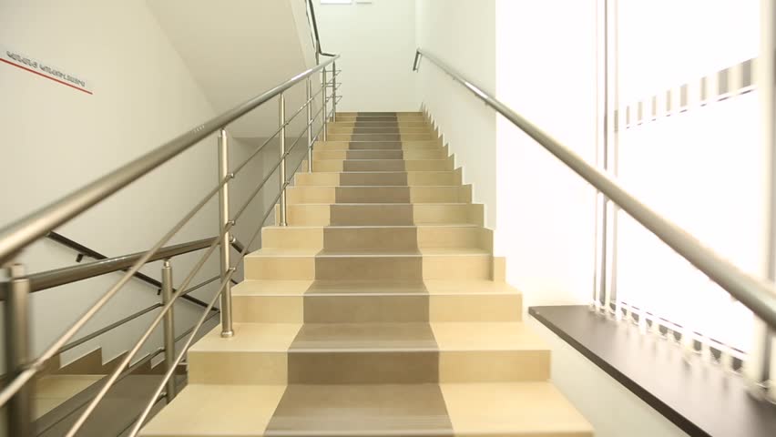 A modern interior staircase with beige and gray tiled steps and sleek metal railings. Sunlight streams in from a window, illuminating the clean, bright space.