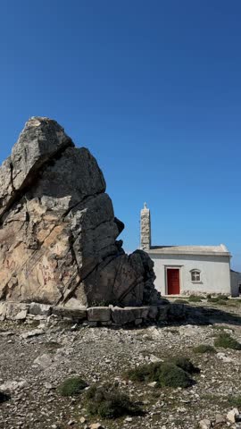 Naxos, Cyclades, Greece, April 18, 2026 – Fanariotisa, a small Orthodox Christian church on a mountainside, partially visible behind a tall natural rock formation under a bright blue sky , with a slow zoom and slight pan movement. 