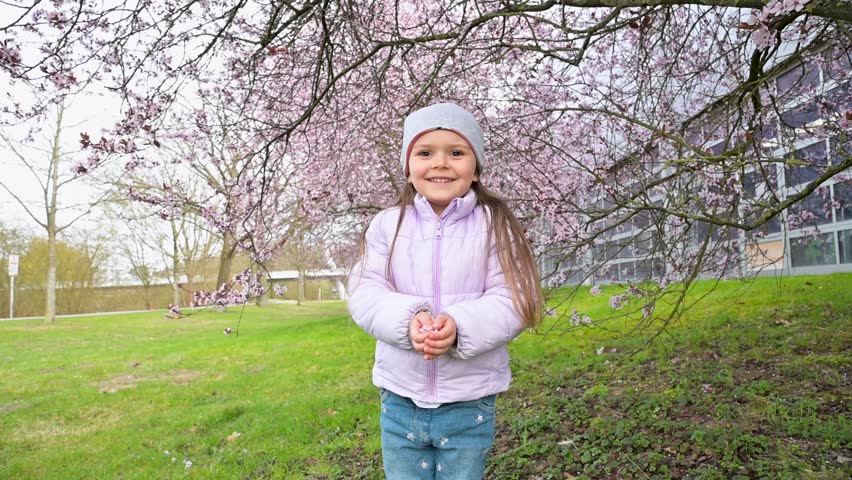 A joyful smiling toddler girl in a spring coat stands under a blooming cherry blossom tree and throws petals into the air in slow motion HD footage