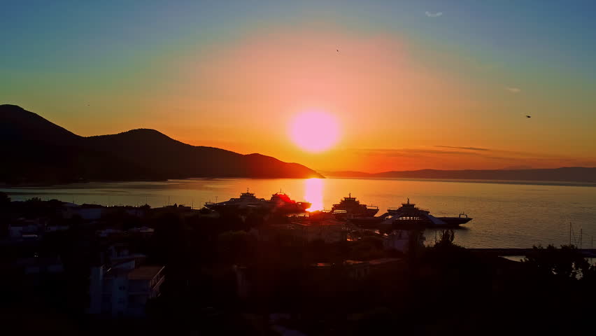 Drone flying forward over ferry port in Thasos Greece at sunset, capturing boats, harbor infrastructure and golden reflection on calm sea with smooth cinematic motion.