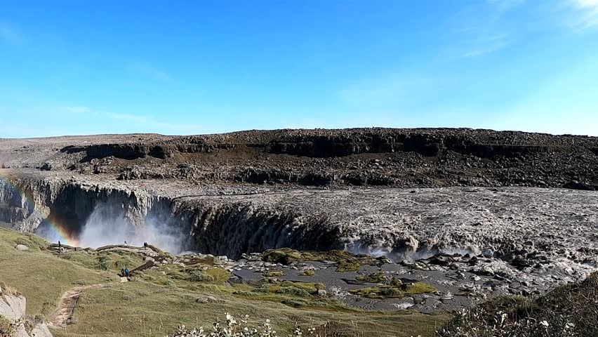 A spectacular wide-angle 4K shot of the mighty Dettifoss waterfall in North Iceland, showcasing the vast rocky landscape of Jökulsárgljúfur canyon. A bright, vivid rainbow is visible in the waterfall