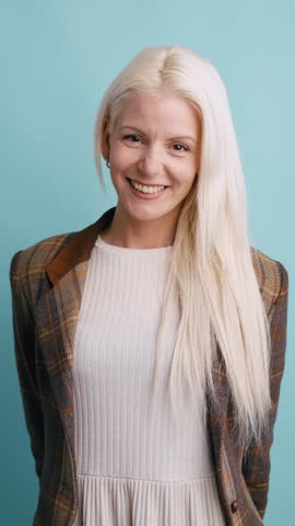 Joyful blonde woman in a stylish plaid blazer smiling at the camera. Her long, beautiful hair is blowing in the wind against a studio background. Vertical