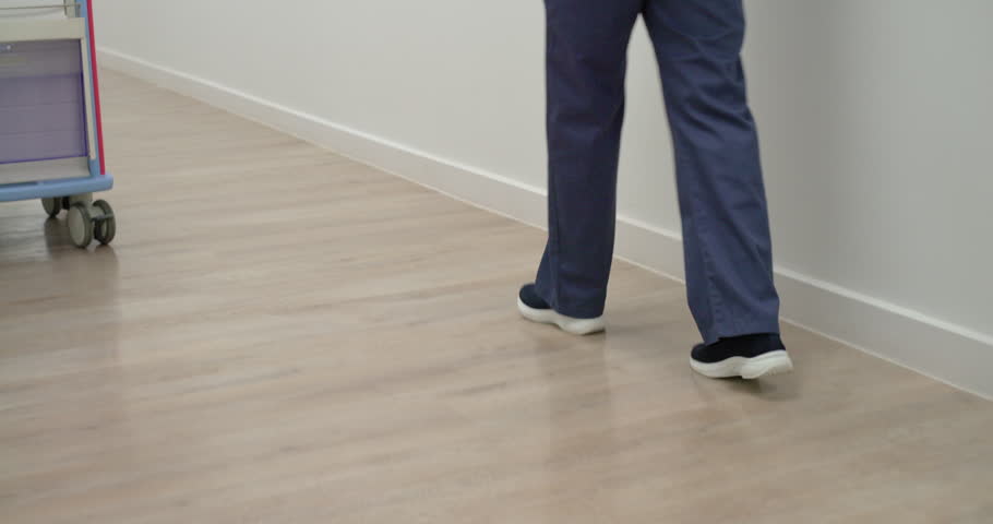 Camera starting low showing nurse in navy scrubs walking with tablet past med cart checking board. Hospital, corridor, clinician, staff, clipboard, modern, clean