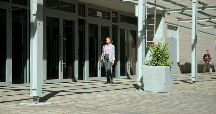Glass doors opening and sending pedestrians moving through plaza, passing planter and posts. Urban, outdoor, crowd, daylight, motion, architecture, shadows
