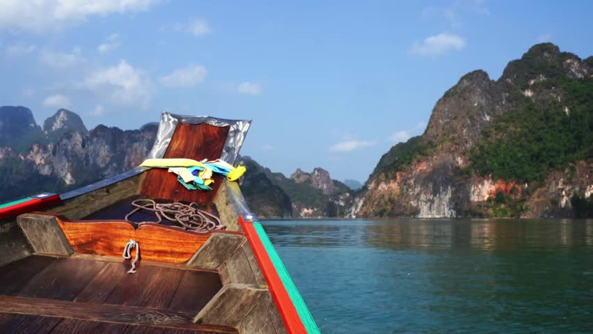 Wooden boat floats calm lake. Green mountains rise behind, steep and proud. Sky is blue, lightly clouded. Water ripples gently, reflecting the view. Peaceful moment, no people in frame