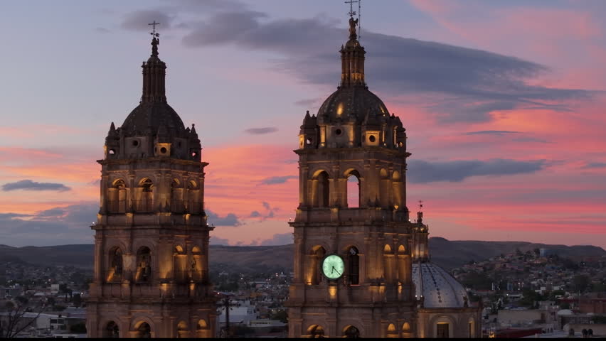 Cathedral Basilica of Immaculate Conception in Durango Mexico towers illuminated against a vibrant pink and orange sunset sky. Colonial baroque architecture with clock tower and crosses over.