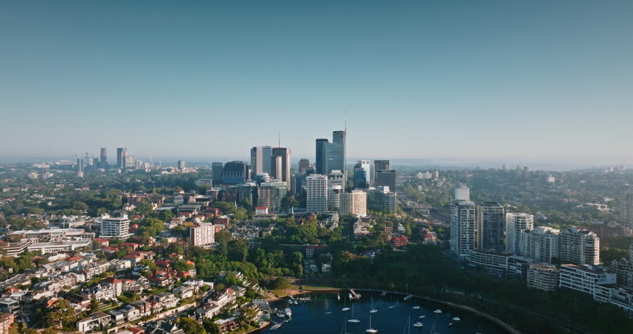 Sydney North skyline rises above a tranquil bay filled with moored boats, surrounded by dense green trees and sprawling residential buildings skyscrapers under clear blue sky. Aerial view drone flight