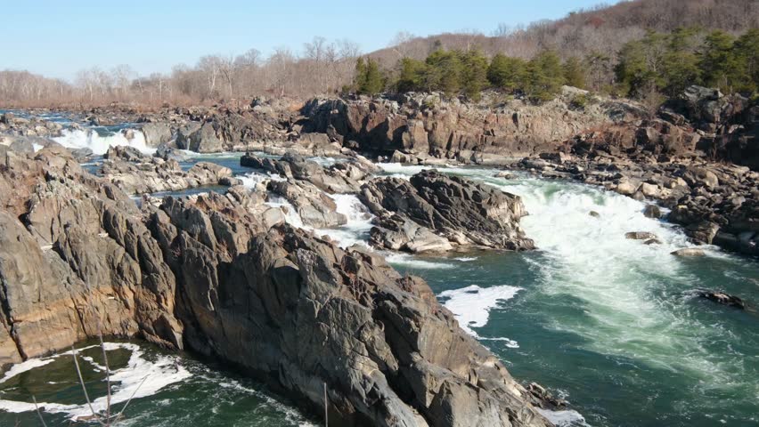 View of Great Falls Park rapids and waterfalls flowing over jagged rocks under a clear blue sky with bare trees on the riverbanks McLean, Virginia, United States.