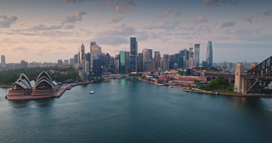 Panoramic view of Sydney sunset cityscape with iconic landmarks - the Opera House and Harbour Bridge, bathed in the soft light of dusk with city skyscrapers in the background. Aerial view drone flight