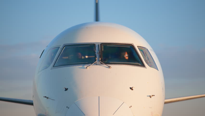 Front view of white commercial aircraft nose section featuring cockpit windows and pitot tubes. Pilots visible inside flight deck during golden hour with soft blue sky background.