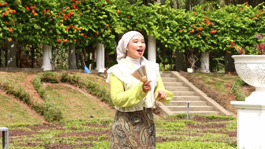 Beautiful young asian woman wearing a hijab smiling and dancing joyfully with a fan in her hand while spending a sunny day outdoors in a beautifully manicured public park