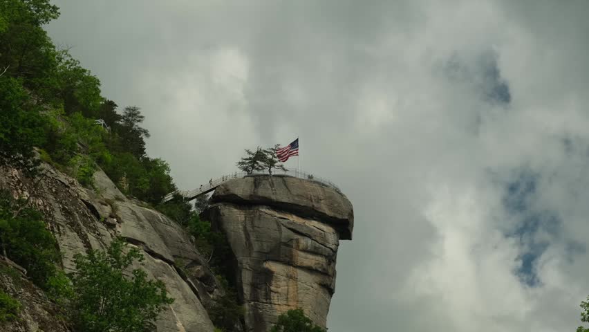 A tall rock formation stands proudly with an American flag atop it. Visitors can be seen around the area, taking in the sights. The sky is cloudy as nature surrounds the site. Chimney Rock