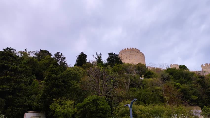 A professional shot of the iconic Rumeli Fortress (Rumeli Hisari) located on the European side of the Bosphorus in Istanbul, Turkey. The medieval Ottoman tower rises majestically from a dense forest of green trees against a dramatic cloudy sky. This high-quality footage captures the grandeur of 15th-century military architecture and historical heritage. Perfect for travel documentaries, educational content, Turkish tourism promotions, and cinematic historical storytelling projects.
