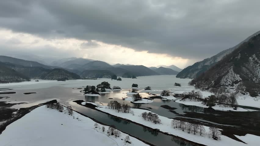 Aerial drone view of a snow-covered mountain rising over Akimoto Lake on a quiet winter morning, Fukushima Prefecture, Japan.