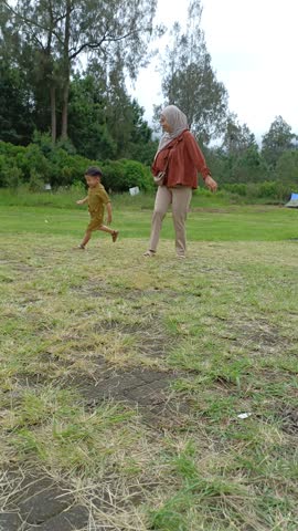 Mother and child run joyfully across a grassy field. They laugh, arms outstretched, enjoying a sunny day outdoors. The child wears a bright yellow outfit, matching their playful energy