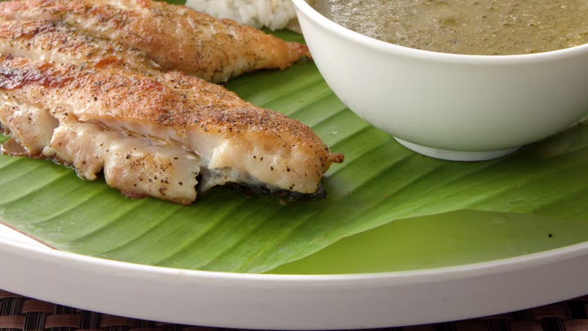 Close up of person serving rice from a large plate of food with grilled fish and fruit.
