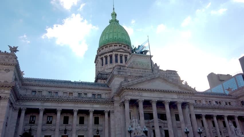 Slow motion low-angle shot of the National Congress of Argentina in Buenos Aires. The majestic neoclassical architecture and green dome stand out against a bright blue sky with clouds