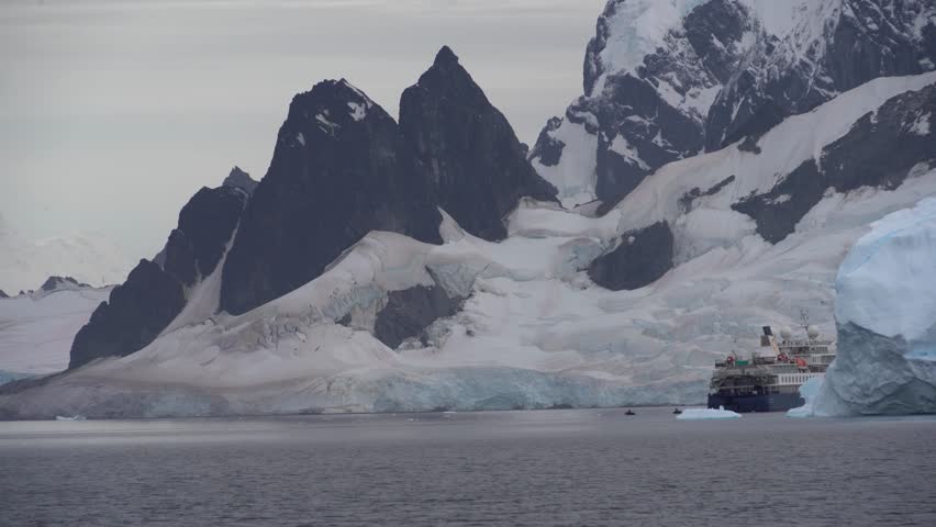 A static shot of a large expedition cruise ship positioned next to a massive iceberg, dwarfed by the towering, dark mountain peaks of the Lemaire Channel in Antarctica