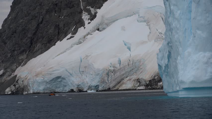 A static shot of a small orange expedition boat dwarfed by a towering blue iceberg and the jagged, snow-covered cliffs of Orne Harbour along the Antarctic Peninsula