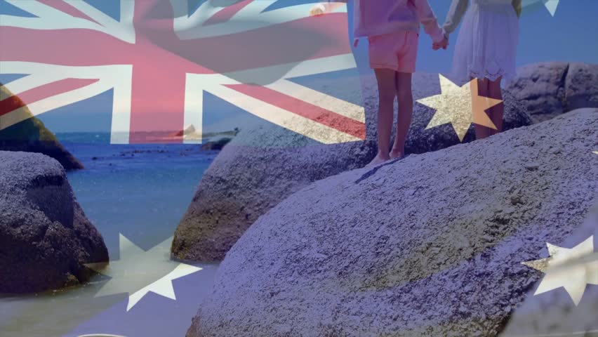 Two girls standing barefoot on rocks holding hands as flag overlay waving across sky for marketing. Children, boulders, coastline, seascape, horizon, stars, unity