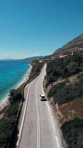 Road Trip in Greece: White Car Navigating a Beautiful Coastal Highway. A white car drives along a winding seaside highway in Greece during a sunny day. This cinematic shot evokes a sense of freedom and adventure, making it ideal for car rental ads, travel vlogs, or Mediterranean lifestyle content.
