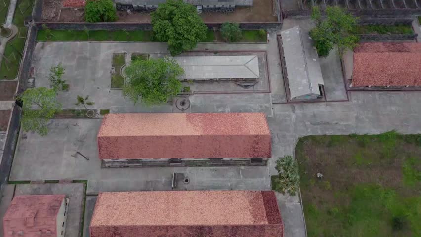 Cinematic aerial drone footage of the historic Tiger Prison complex on Con Dao island, Vung Tau province, southern Vietnam. The top-down perspective reveals the austere colonial-era prison architecture — rows of long barrack buildings with terracotta tiled roofs arranged within high perimeter walls, surrounded by tropical vegetation and open courtyards. Con Dao