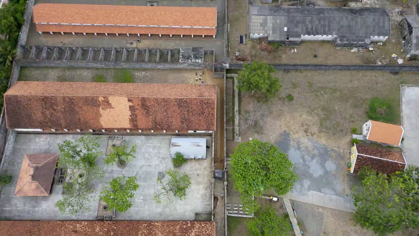 Cinematic top-down aerial drone footage flying over the historic Tiger Prison compound on Con Dao island, Vung Tau province, southern Vietnam. The camera looks directly down revealing the interior layout of the colonial-era prison — long barrack buildings with weathered terracotta tiled roofs arranged around open courtyards, surrounded by high perimeter walls. The austere architecture shows visible signs of age and deterioration, with moss-covered roofs and overgrown courtyard trees adding to the somber atmosphere. Con Dao