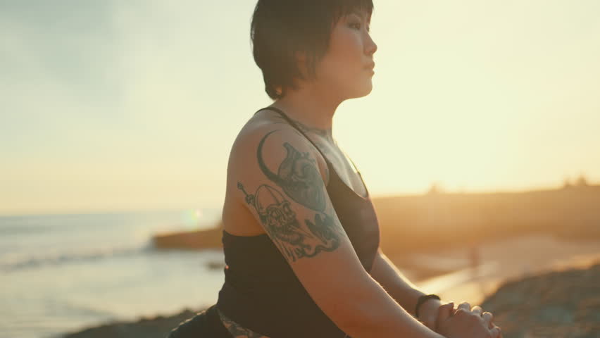 Woman with tattoos stretching her legs during a workout on a cliffside by the sea at sunrise