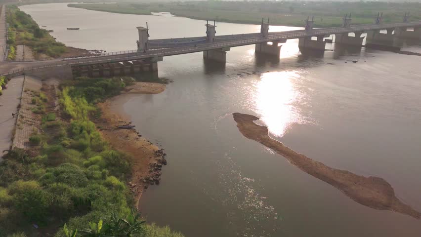 Beschrijving:
Cinematic aerial drone footage of the Tra Khuc river barrage spanning the wide muddy river in Quang Ngai city, central Vietnam. The camera reveals the full structure of the hydraulic dam and road bridge combination — multiple concrete gate pillars and sluice gates controlling the flow, with a narrow sandbar emerging from the low water level downstream. The late afternoon sun reflects off the calm brown river surface, creating a warm golden shimmer. Lush riverbank vegetation and flat agricultural land frame both sides of the river. A traditional boat is faintly visible upstream. Shot in 4K UHD. Ideal for infrastructure, engineering, water management, rural Vietnam, and aerial landscape documentary content.