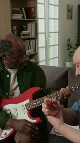 Vertical shot of focused senior African American man learning to play electric guitar with male music teacher helping and adjusting his hand position Useful for educational services