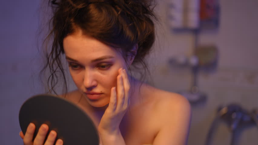 Young thoughtful woman wrapped in a towel looking at her reflection in a hand mirror, examining her face for imperfections after a shower