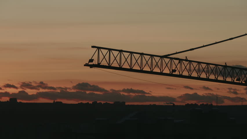 Silhouette of a large tower crane operating over a city skyline against the warm, dramatic colors of the setting sun, with dark clouds moving slowly across the orange and red sky. timelapse