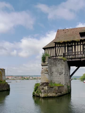 Vernon, France - May 23 2023: Half-timbered structure known as the Old Mill stands on a broken bridge over the Seine River in Vernon, Normandy, France. The building, characterized by its timber beams and tiled roof, straddles two stone sections of the bridge. Boats are visible on the river beneath it, surrounded by lush greenery on the riverbanks. The sky is partly cloudy, providing a picturesque backdrop to the historic structure.
