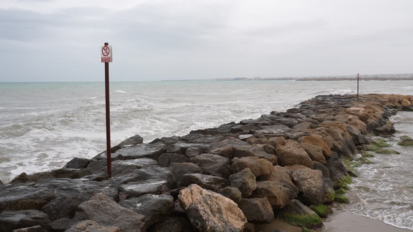 Stormy sea with waves crashing against rocky breakwater. Warning sign on pier. Cloudy weather, dramatic coastal scene in Mediterranean urban shoreline.