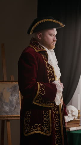 Vertical portrait of aristocratic young Caucasian man adjusting tricorn hat wearing embroidered coat and looking at camera