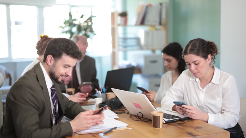 Concentrated office workers scrolling smartphone together sitting at workplace in bright office of business company 