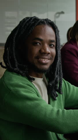 Vertical portrait shot of young African American man with dreadlocks sitting in office and posing for camera with friendly smile
