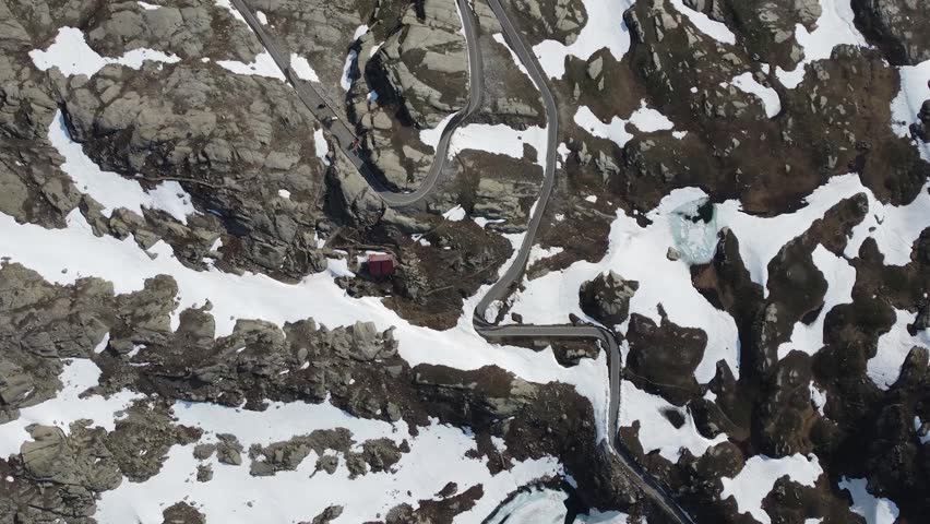 Aerial view of serpentine road winds through a rugged, snow-dusted mountain landscape, contrasting with a small red cabin, Aosta, Aosta Valley, Italy.