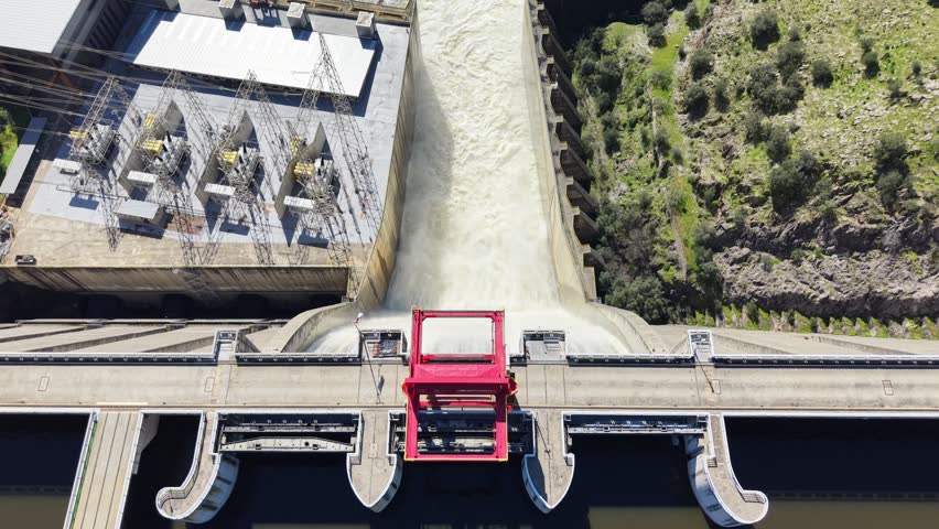 Aerial view of a powerful hydroelectric dam releasing torrents of foamy white water, contrasting with rugged green hills, Alcántara, Extremadura, Spain.