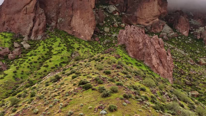 Aerial view of imposing reddish-brown rock formations emerging from a misty cloud, contrasting with vibrant green vegetation on the slopes, Apache Junction, Arizona, United States.
