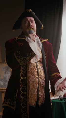 Vertical shot of Caucasian man wearing embroidered coat, tricorn hat and lace cravat singing into microphone with passion indoors