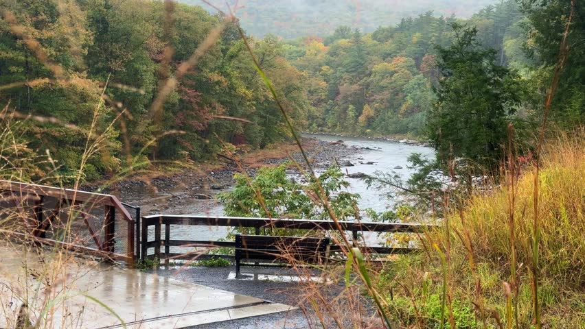 Rainy autumn overlook above a rocky river in the Catskill Mountains, with a footbridge, wet foliage, and misty forested hills under a moody Appalachian sky.