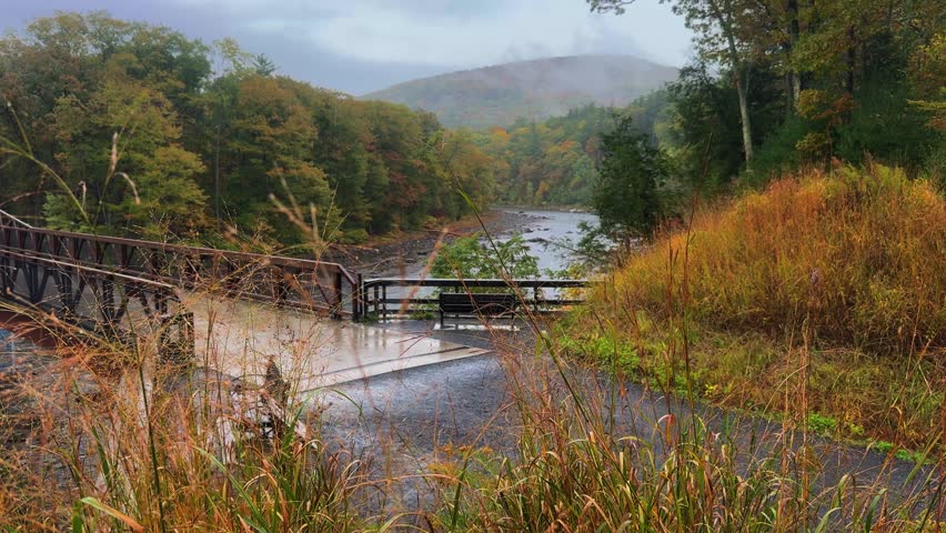 Foggy, rainy autumn overlook in the Catskill Mountains, featuring a footbridge and bench above a river, wet foliage, and misty forest hills moving in the breeze.