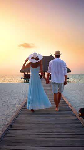 A romantic holiday couple walks down a wooden pier in the Maldives islands during sunset time