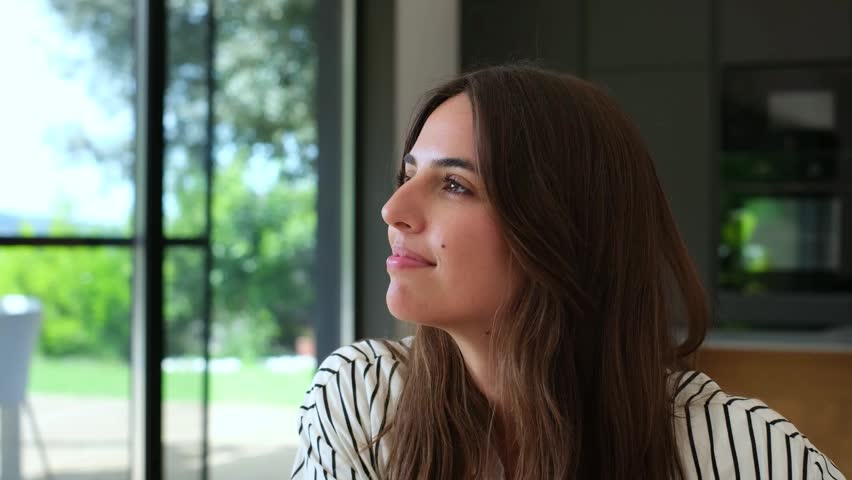 A beautiful young woman smiles softly while looking out a large window in a modern home, embodying feelings of contentment and happiness.