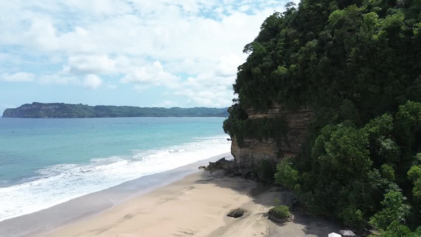 Cinematic drone view of a pristine seaside landscape, featuring gentle waves breaking on the shore and a massive forested headland.