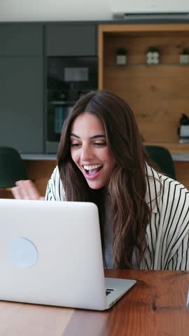 A happy young woman engages in a lively video call on her laptop, smiling and gesturing from a modern home office. Her expressive face conveys joy and excitement during the virtual communication.