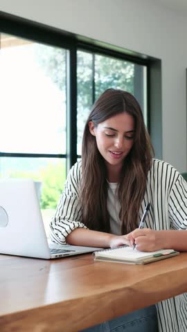 A happy young woman works diligently from her home office, smiling and taking notes in a notebook next to her laptop. She enjoys a bright, comfortable, and productive workspace.